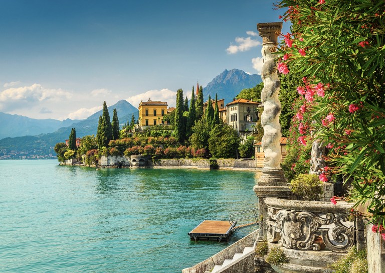 Scenic view of villas by a lake with mountains, greenery, and flowers.